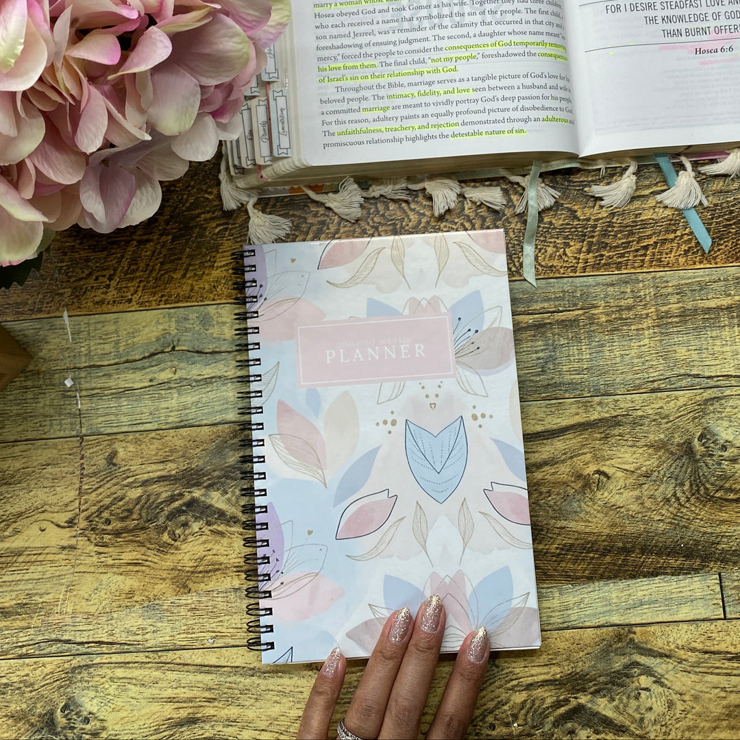A styled flat-lay on a rustic wooden table featuring a small spiral-bound notebook with a pink floral cover on the left, held by a hand, and an open book with highlighted pages on the right next to a large pink hydrangea.