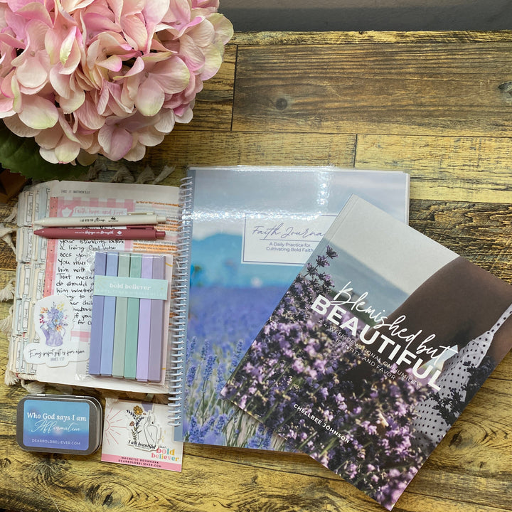 A styled flat-lay of stationery and journaling supplies on a rustic wooden table, including a spiral notebook, a planner pad, pastel sticky notes, a pink pen, a small blue tin, and a floral print card, next to a large pink hydrangea.