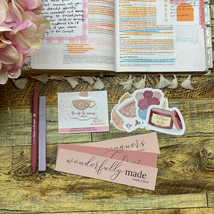 An open handmade journal with colorful written and illustrated pages, styled on a rustic wooden table alongside pink paper tags, stickers, a pink pen, and a large pink flower.