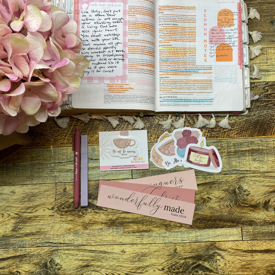 A styled flat-lay of a pink and white planner sticker set on a rustic wooden table next to an open journal and a pink hydrangea. The stickers include cute illustrations of a cupcake, a heart, and a small character, with one sticker reading 'whispers made'.