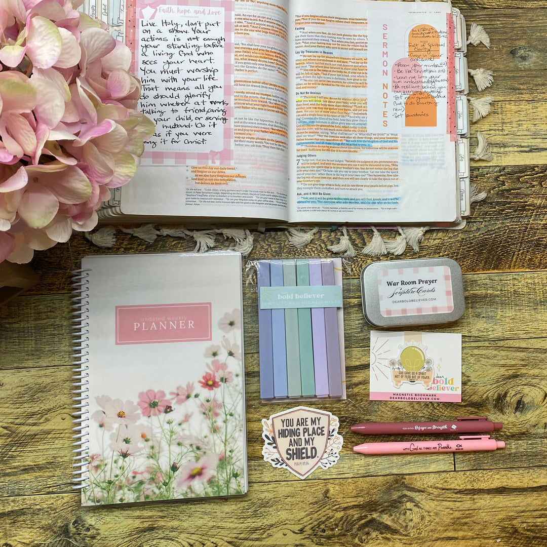A styled flat-lay of stationery items on a rustic wooden tabletop, including a floral planner, an open notebook with handwritten pages, pink pens, a small card, a sticker, and a pink hydrangea flower.