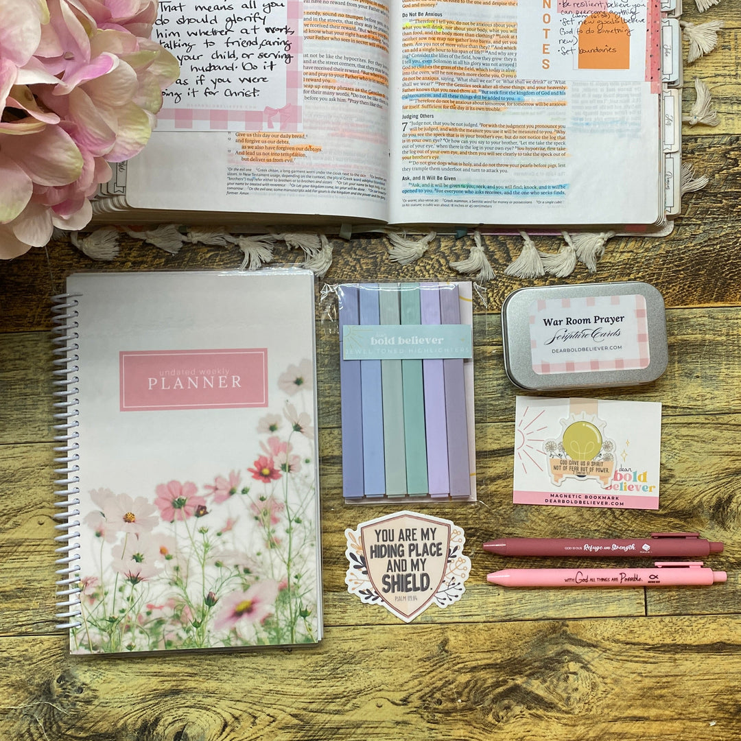 A styled flat-lay of stationery items on a rustic wooden table, including a floral planner, an open notebook with colorful page markers, pink pens, a stack of pastel sticky notes, small cards, and a pink hydrangea flower arrangement.