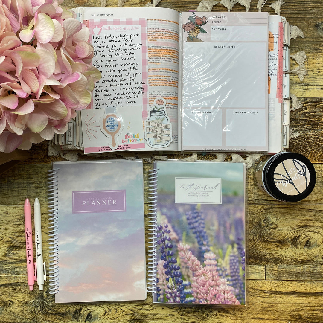 A styled flat-lay of stationery items on a rustic wooden table, including a closed pastel planner, a closed floral planner, an open planner with handwritten notes, pink pens, a small round black item, and a large pink flower arrangement.