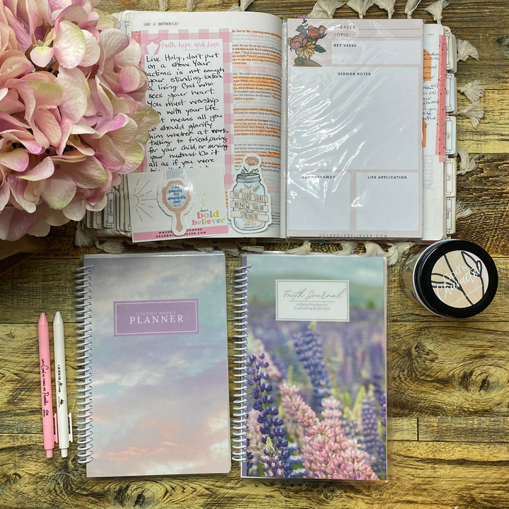 A styled flat-lay of stationery items on a rustic wooden table, including a closed pastel planner, a closed floral planner, an open planner with handwritten notes, pink pens, a small round black item, and a large pink flower arrangement.