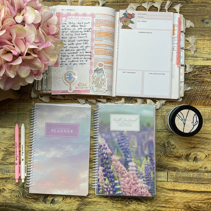 A styled flat-lay of stationery items on a rustic wooden table, including a pastel iridescent notebook, a lavender floral notebook, an open planner with handwritten notes, pink pens, a small round black item, and a large pink hydrangea flower.