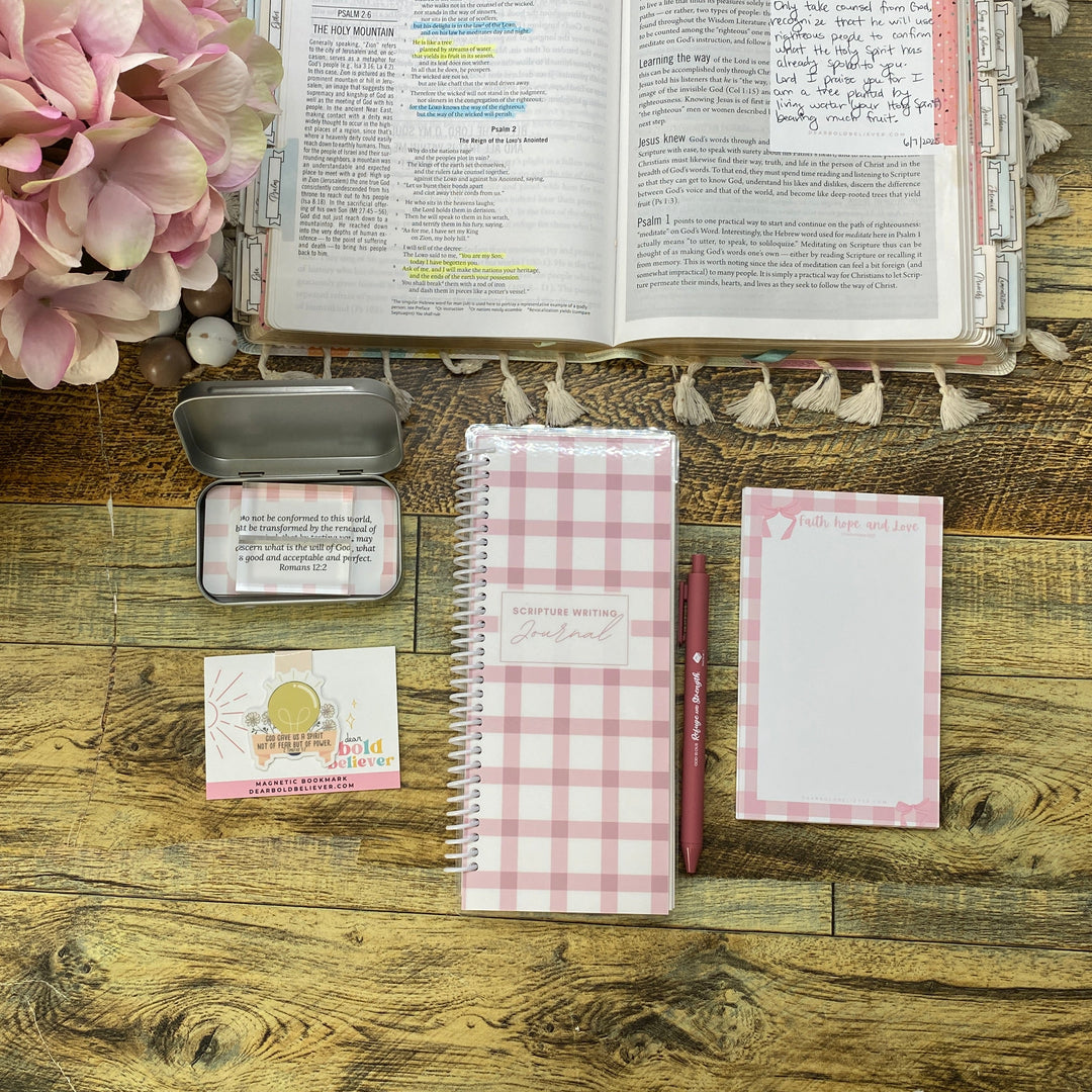 A styled flat-lay of stationery items on a rustic wooden table, including a pink and white gingham spiral notebook, a matching notepad, a small metal tin, a card, and an open book, with pink flowers in the upper right corner.