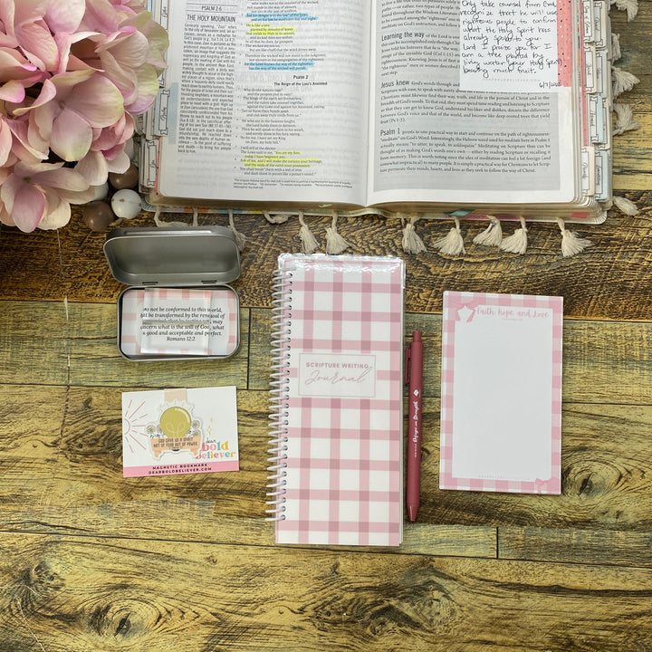 A styled flat-lay of stationery items on a rustic wooden table, including a pink and white gingham spiral notebook, a matching notepad, a small metal tin, a card, and an open book, with pink flowers in the upper right corner.