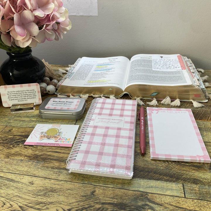 A styled desk setup featuring a pink and white gingham spiral notebook, a matching notepad, and a pink pen, arranged on a rustic wooden table alongside an open Bible, a small sign, and a vase of pink flowers.