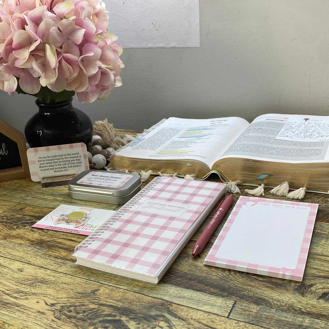 A styled desk setup featuring a pink and white gingham notepad, a matching pink pen, and a blank notepad on a rustic wooden table, with an open Bible, a vase of pink flowers, and a small chalkboard sign in the background.