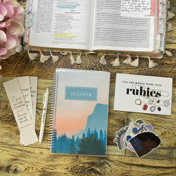 A styled flat-lay of a spiral-bound planner on a rustic wooden table, surrounded by decorative pink flowers, an open notebook, cards, and stickers. The planner cover features a scenic mountain and forest illustration with the word 'PLANNER' in a blue box.
