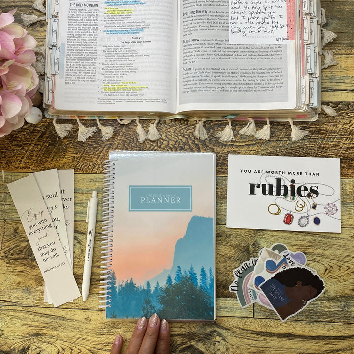 A styled desk flat-lay featuring a spiral-bound planner with a mountain landscape cover, an open Bible, a pink floral arrangement, stickers, and a hand resting on the left side of the wooden table.