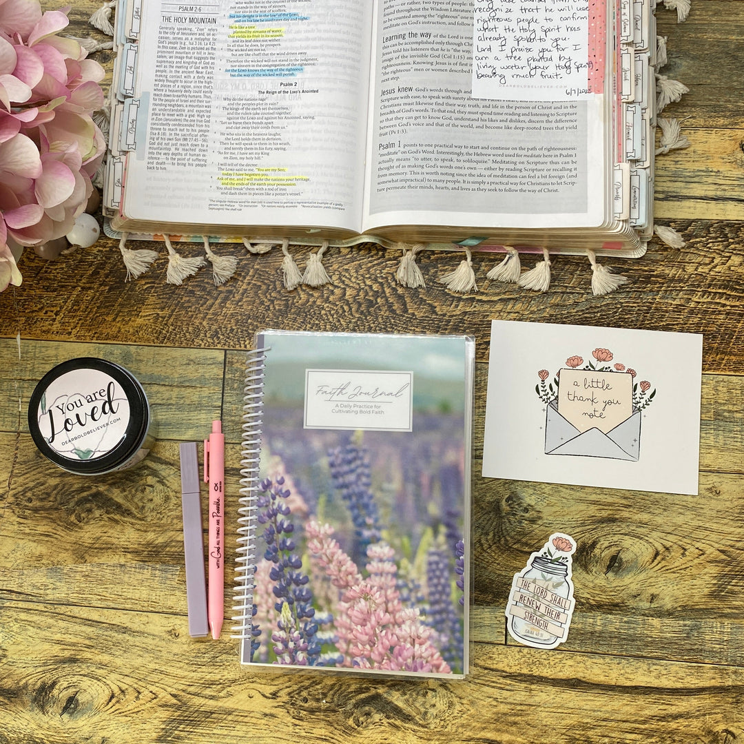A styled desk flat-lay featuring a floral notepad, a pink pen, a small round tin, a sticker, a postcard, and an open planner or book on a rustic wooden table next to a pink flower arrangement.