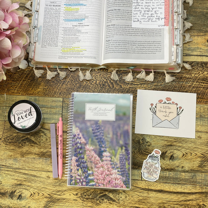 A styled desk flat-lay featuring a floral notepad, a pink pen, a small round tin, a sticker, a postcard, and an open planner or book on a rustic wooden table next to a pink flower arrangement.