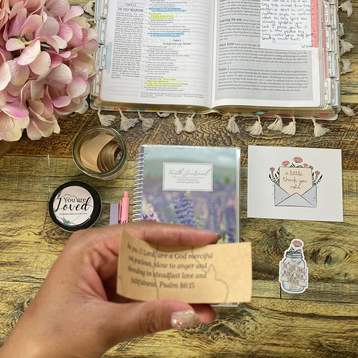 A hand holds a small kraft paper tag with printed text over a rustic wooden desk. The desk is styled with a pink hydrangea, an open book, a small notebook, a jar, and other stationery items.