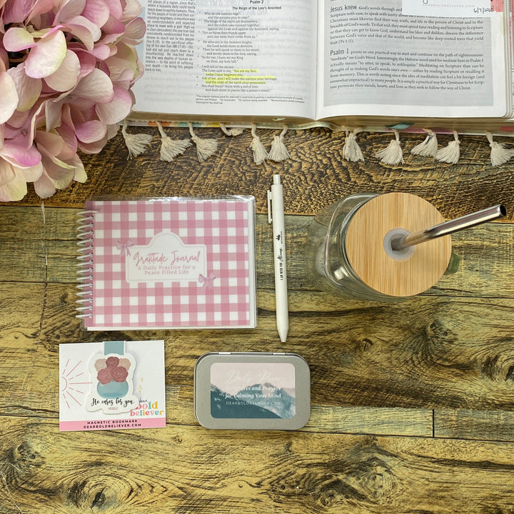 A styled tabletop arrangement on a rustic wood surface featuring a pink hydrangea, an open Bible, a pink-and-white gingham spiral notebook, a white pen, a glass jar with a bamboo lid and metal straw, a small card, and a small rectangular tin.
