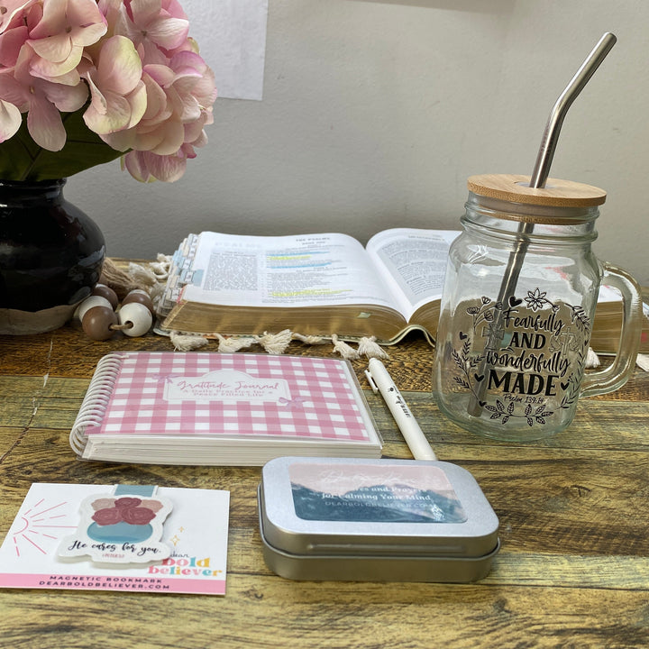 A styled tabletop arrangement featuring a pink gingham notebook, a silver metal tin, a pink and white card, and a glass mason jar with a metal straw on a rustic wooden table.