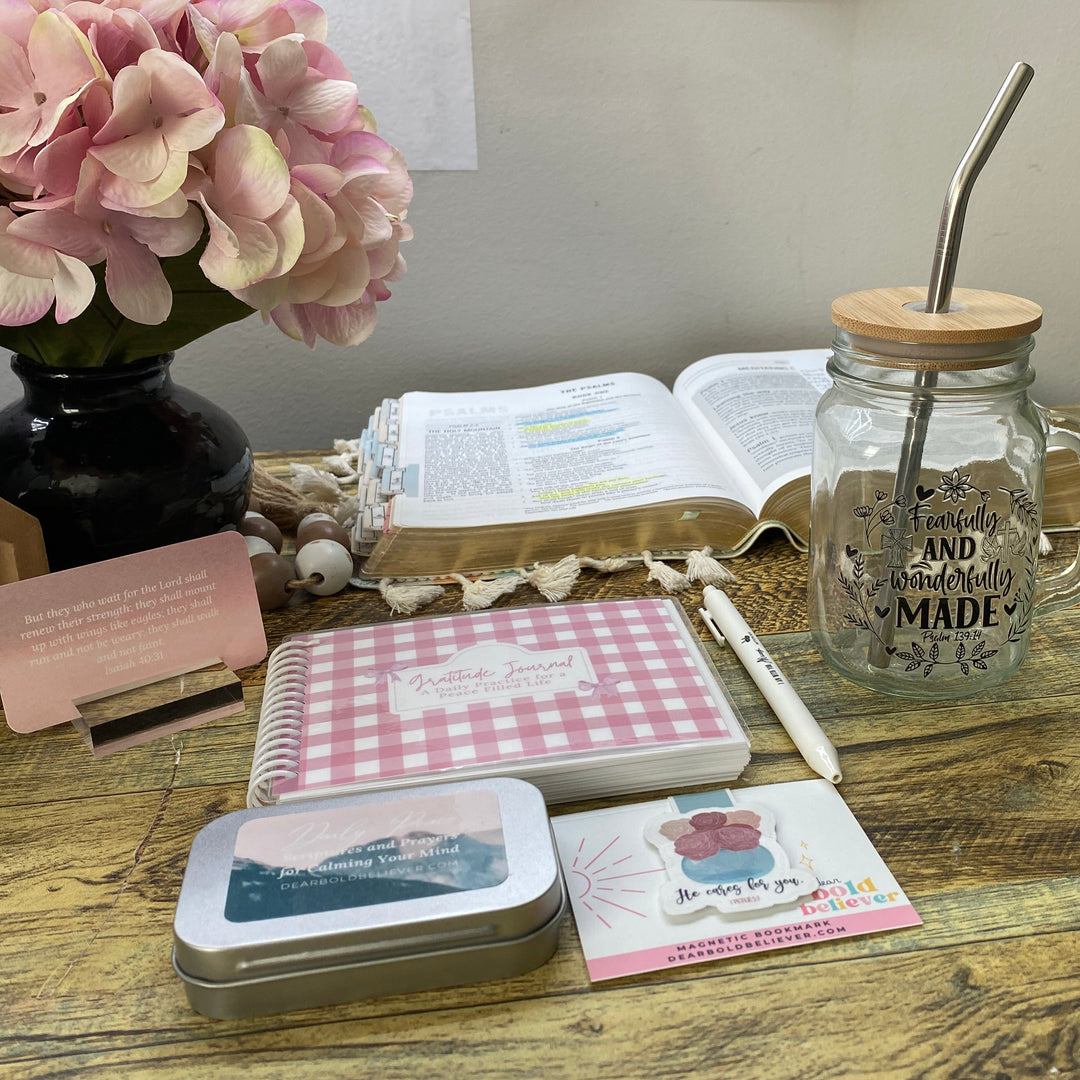 A styled tabletop display featuring a pink gingham notebook, a small metal tin, a card, a pen, an open book, and a glass mason jar with a metal straw on a rustic wooden table.