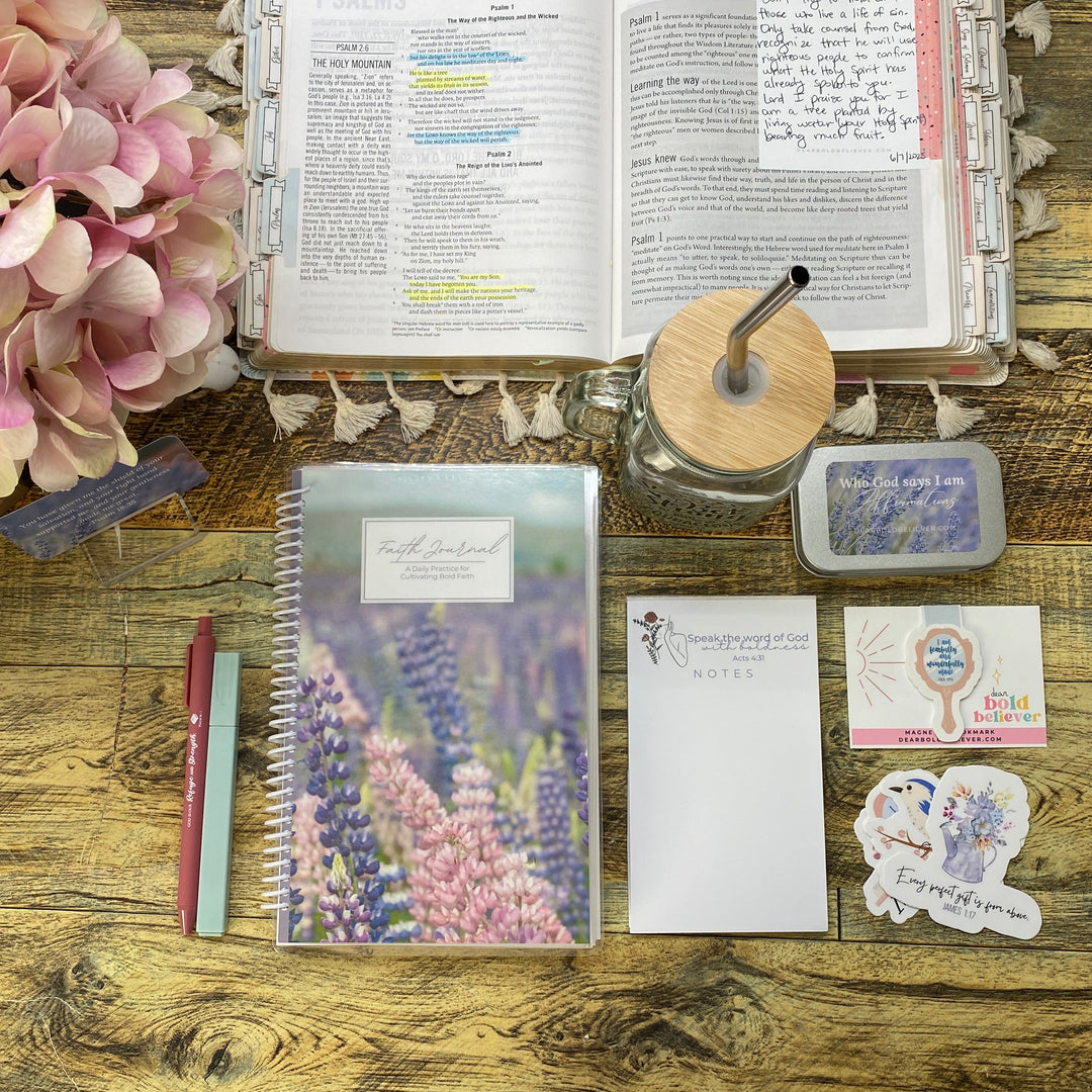 A styled desk flat-lay featuring a floral purple and pink spiral notebook, a matching notepad, stickers, a pen, a glass jar with a wooden lid, and an open Bible on a rustic wooden table.