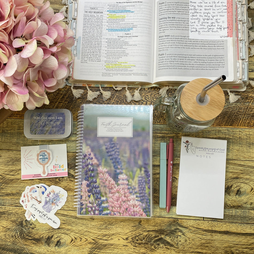 A styled desk setup featuring a lavender and pink floral spiral notebook, an open Bible with sticky notes, a glass jar with a wooden lid, a small container of lavender buds, and decorative stickers on a rustic wooden table next to a pink hydrangea.