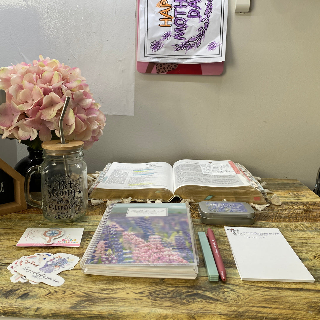A styled Mother's Day desk setup on a wooden table, featuring a glass mason jar filled with pink flowers, an open Bible, a floral planner, a small chalkboard sign, stickers, and notepad.