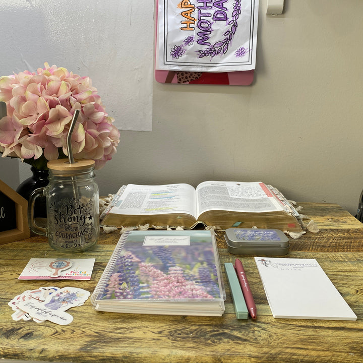 A styled Mother's Day desk setup on a wooden table, featuring a glass mason jar filled with pink flowers, an open Bible, a floral planner, a small chalkboard sign, stickers, and notepad.