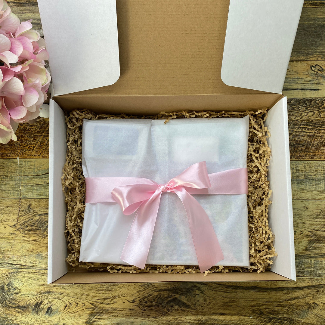 A white gift box filled with brown crinkle paper packing material, containing a white-wrapped item tied with a light pink satin ribbon. The box sits on a rustic wooden tabletop next to pink flowers.