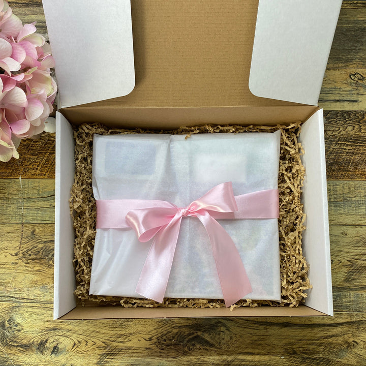 A white gift box filled with brown crinkle paper packing material, containing a white-wrapped item tied with a light pink satin ribbon. The box sits on a rustic wooden tabletop next to pink flowers.