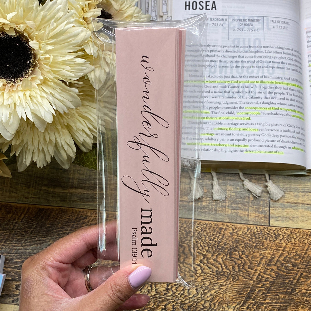 Pink 'Wonderfully Made' bookmark held by a hand with flowers and an open book in the background.