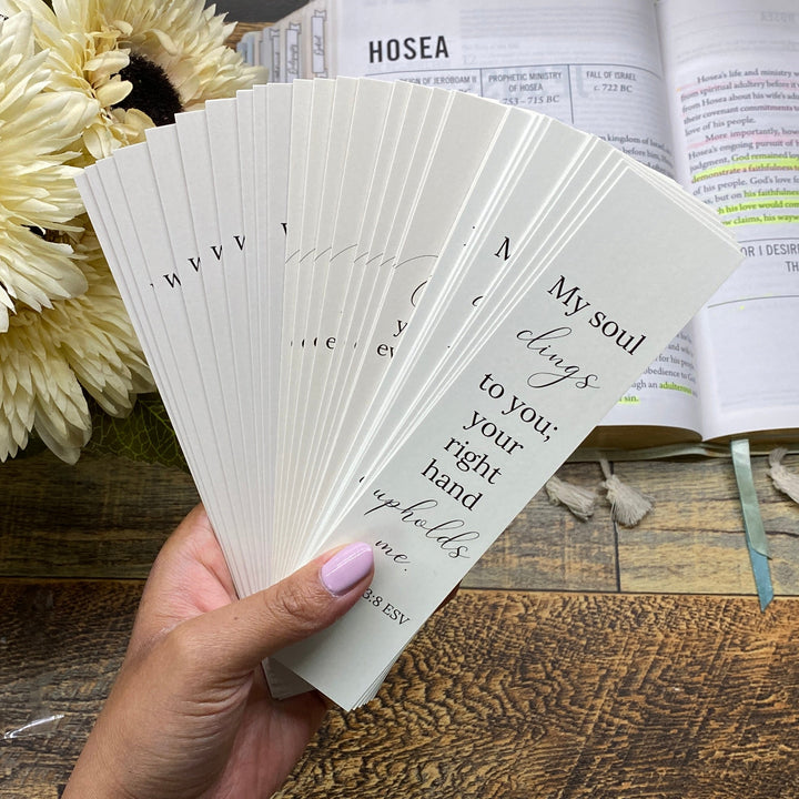 Hand holding prayer cards in front of an open Bible on a wooden surface with flowers.
