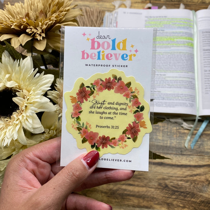 Hand holding a card with floral design and text, placed on a wooden surface with flowers.