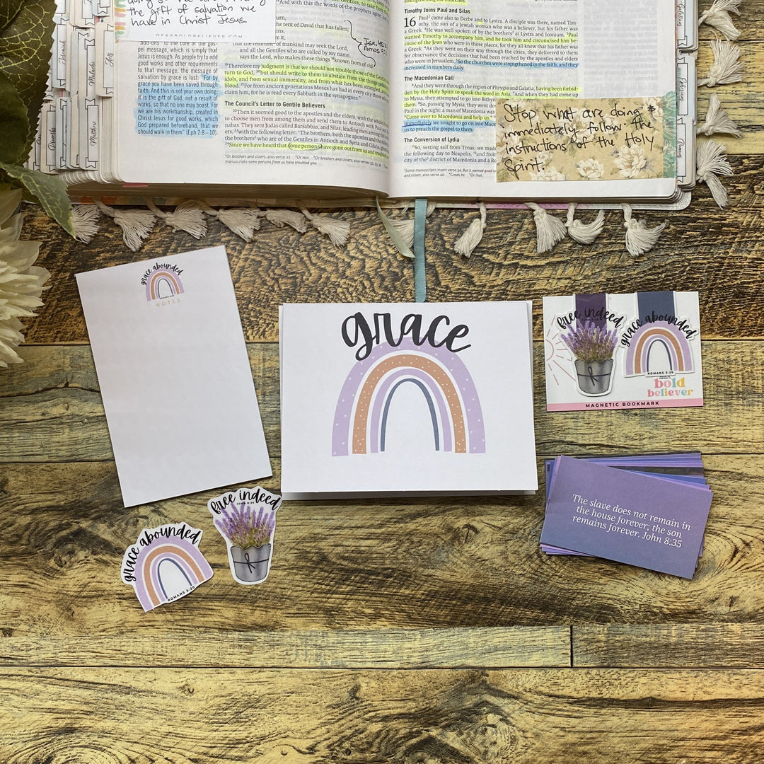 A coordinated stationery set arranged on a rustic wooden surface, including a white card with a pastel rainbow and the word 'grace,' matching envelopes, small die-cut stickers, and a purple card. An open book and dried flowers are visible in the background as styled props.