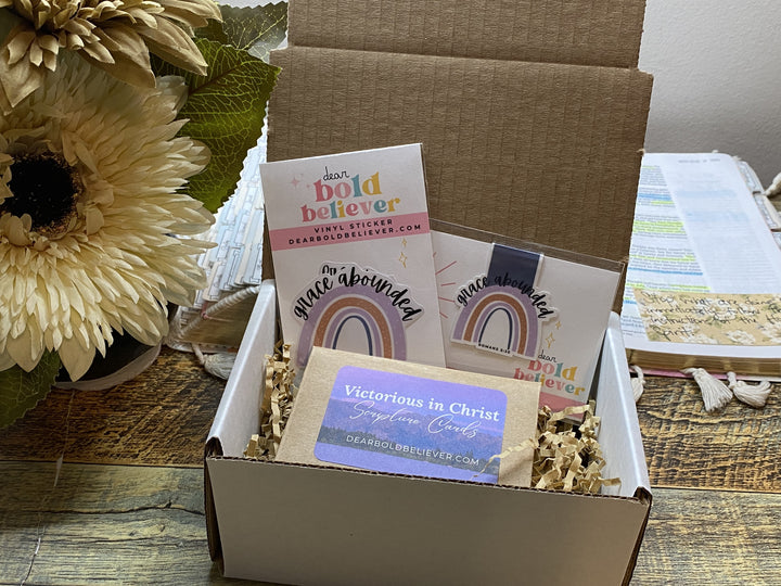 An open kraft cardboard gift box filled with prayer cards and paper shred, placed on a wooden surface next to large cream flowers. Visible card text includes 'damn bold believer', 'peace abounded', 'Victorious in Christ', and 'grace abounded'.